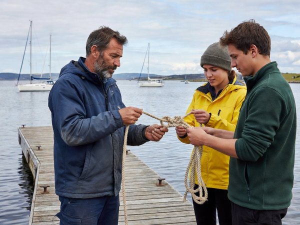 A sailing instructor demonstrating a bowline knot to two students on a pontoon